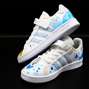 K&ouml;nnte beinhalten: Ein Paar wei&szlig;e Adidas Sneaker mit blauen und wei&szlig;en Designs, inspiriert vom Film Frozen. Ein Schuh zeigt ein Schneeflockenmuster und der andere zeigt Olaf, den Schneemann.