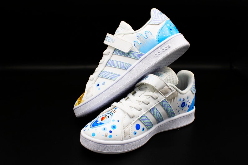 K&ouml;nnte beinhalten: Ein Paar wei&szlig;e Adidas Sneaker mit blauen und wei&szlig;en Designs, inspiriert vom Film Frozen. Ein Schuh zeigt ein Schneeflockenmuster und der andere zeigt Olaf, den Schneemann.