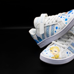 K&ouml;nnte beinhalten: Wei&szlig;e Sneaker mit einem blauen und wei&szlig;en Frozen-Design. Die Sneaker haben einen Klettverschluss mit dem Wort "Frozen" darauf. Das Design zeigt Elsa und Olaf.