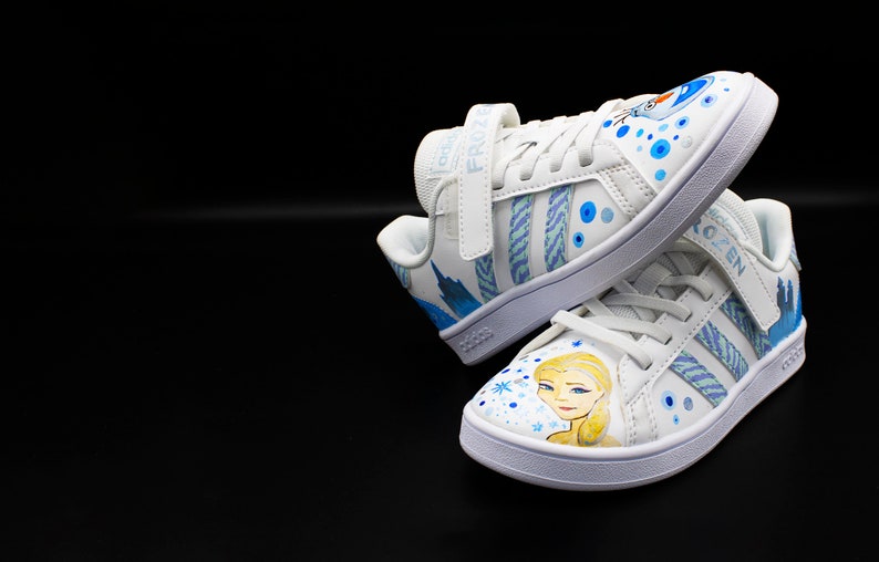 K&ouml;nnte beinhalten: Wei&szlig;e Sneaker mit einem blauen und wei&szlig;en Frozen-Design. Die Sneaker haben einen Klettverschluss mit dem Wort "Frozen" darauf. Das Design zeigt Elsa und Olaf.