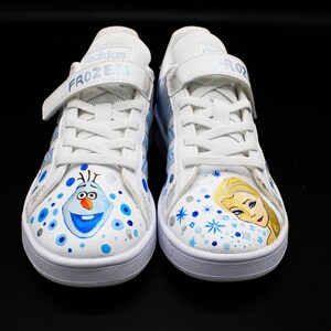 K&ouml;nnte beinhalten: Ein Paar wei&szlig;e Canvas-Sneaker mit blauen und silbernen Akzenten. Ein Schuh zeigt ein gemaltes Bild von Olaf aus Frozen, der andere ein gemaltes Bild von Elsa aus Frozen. Die Schuhe haben Klettverschl&uuml;sse und der Text "FROZEN" ist auf dem Riemen aufgedruckt.