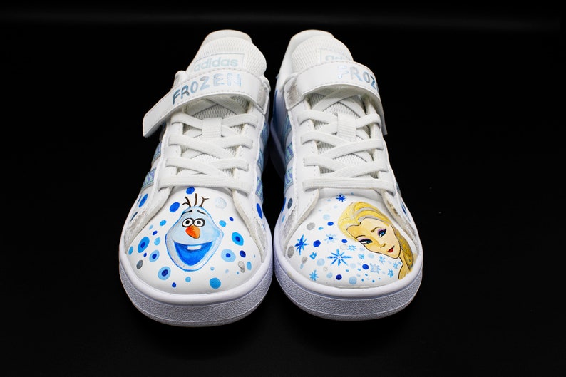 K&ouml;nnte beinhalten: Ein Paar wei&szlig;e Canvas-Sneaker mit blauen und silbernen Akzenten. Ein Schuh zeigt ein gemaltes Bild von Olaf aus Frozen, der andere ein gemaltes Bild von Elsa aus Frozen. Die Schuhe haben Klettverschl&uuml;sse und der Text "FROZEN" ist auf dem Riemen aufgedruckt.