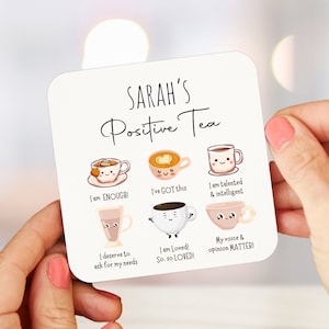 Op de afbeelding: Witte onderzetter met de tekst "SARAH'S Positive Tea" en zes geillustreerde theekopjes met positieve affirmaties. De theekopjes zijn versierd met lachende gezichten en harten. De affirmaties zijn: "I am ENOUGH!", "I've GOT this", "I am talented & intelligent", "I deserve to ask for my needs", "I am Loved! So. so LOVED!" en "My voice & opinion MATTER!"