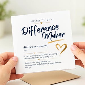 May include: A white greeting card with the words "Difference Maker" in navy blue script and gold accents. The card includes a definition and a gold heart illustration. The card is held by two hands.