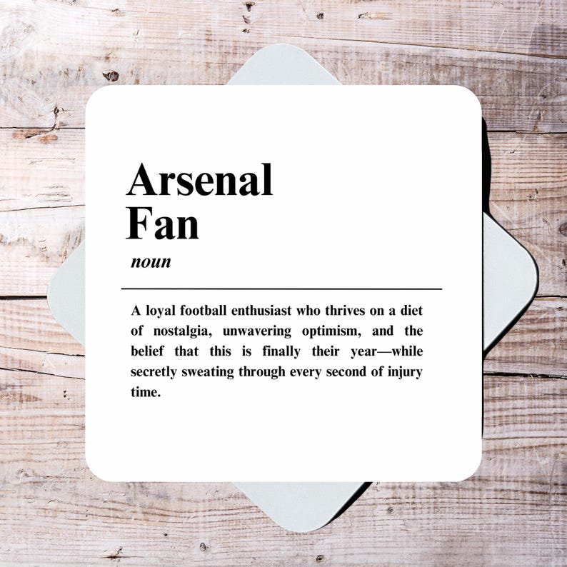May include: A white coaster with black text that reads "Arsenal Fan noun A loyal football enthusiast who thrives on a diet of nostalgia, unwavering optimism, and the belief that this is finally their year-while secretly sweating through every second of injury time."