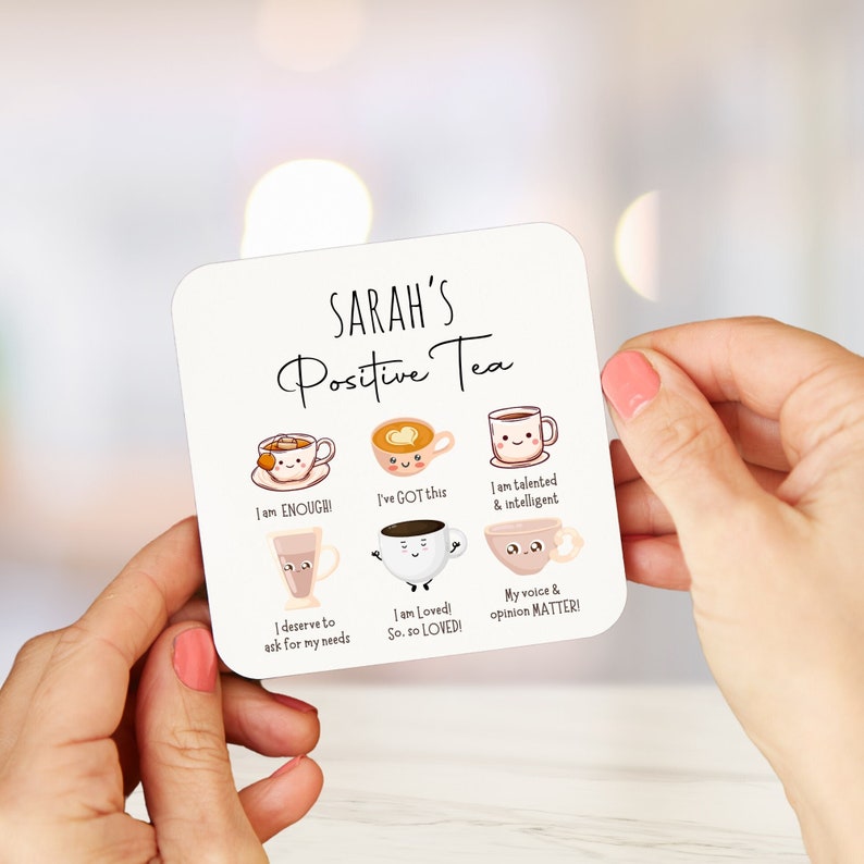 Op de afbeelding: Witte onderzetter met de tekst "SARAH'S Positive Tea" en zes geillustreerde theekopjes met positieve affirmaties. De theekopjes zijn versierd met lachende gezichten en harten. De affirmaties zijn: "I am ENOUGH!", "I've GOT this", "I am talented & intelligent", "I deserve to ask for my needs", "I am Loved! So. so LOVED!" en "My voice & opinion MATTER!"