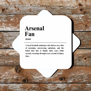 May include: A white coaster with a black and white definition of an Arsenal fan. The definition reads: "A loyal football enthusiast who thrives on a diet of nostalgia, unwavering optimism, and the belief that this is finally their year-while secretly sweating through every second of injury time."