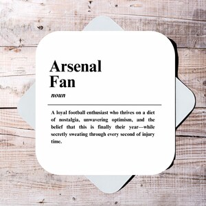 May include: White coaster with black text defining an Arsenal fan as a loyal football enthusiast who thrives on nostalgia, unwavering optimism, and the belief that this is finally their year, while secretly sweating through every second of injury time.