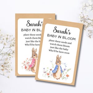 May include: Two seed packet cards with a watercolour illustration of a bunny wearing a blue jacket and pink scarf. The text on the cards reads "Sarah's BABY IN BLOOM" and a message about planting seeds. The cards are on a white background.