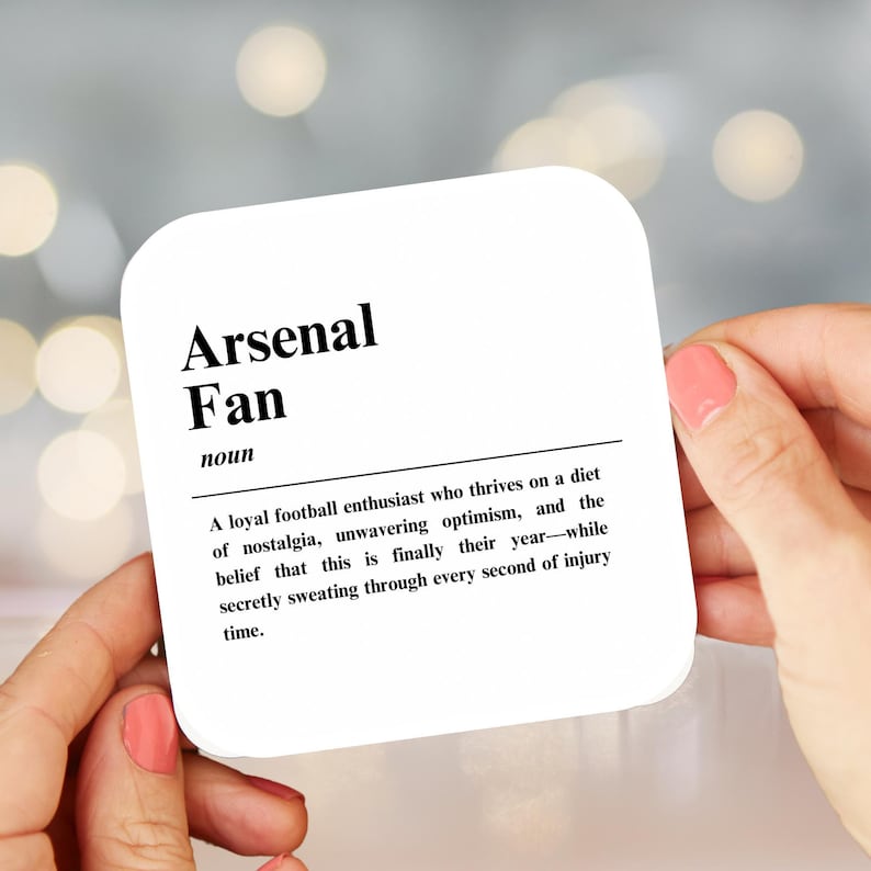 May include: A white coaster with black text that reads "Arsenal Fan" and defines the term as "A loyal football enthusiast who thrives on a diet of nostalgia, unwavering optimism, and the belief that this is finally their year-while secretly sweating through every second of injury time."