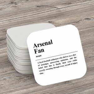 May include: White square coaster with black text that reads "Arsenal Fan" and a definition of the term. The definition reads: "A loyal football enthusiast who thrives on a diet of nostalgia, unwavering optimism, and the belief that this is finally their year - while secretly sweating through every second of injury time."
