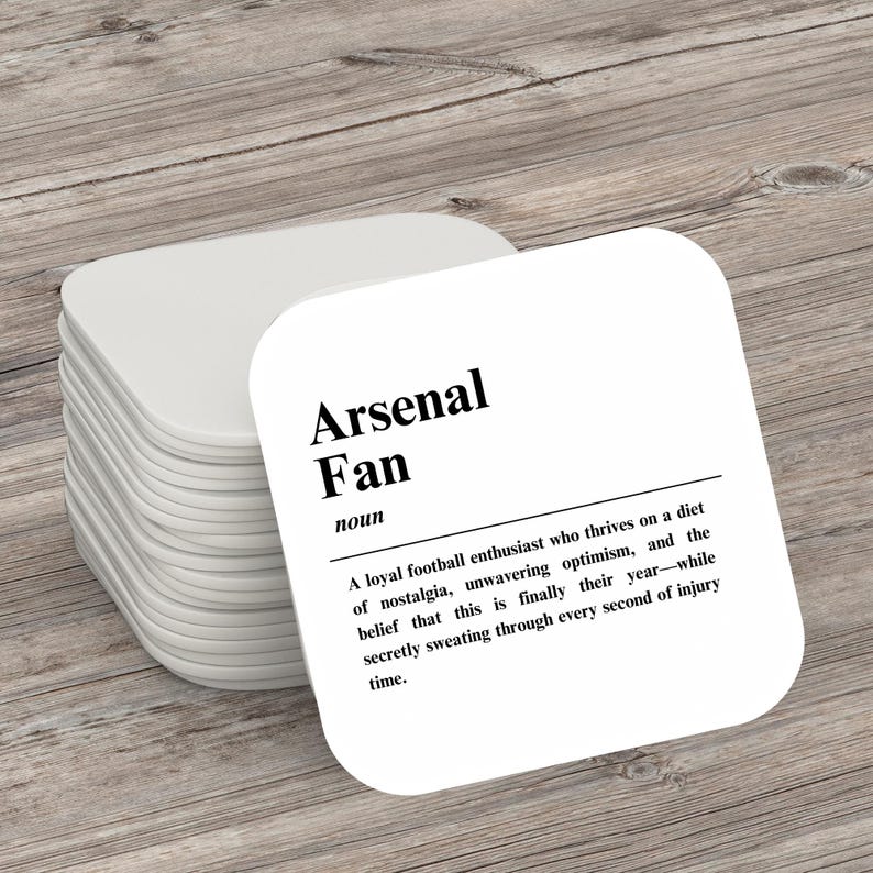 May include: White square coaster with black text that reads "Arsenal Fan" and a definition of the term. The definition reads: "A loyal football enthusiast who thrives on a diet of nostalgia, unwavering optimism, and the belief that this is finally their year - while secretly sweating through every second of injury time."