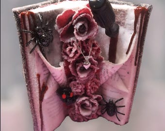 May include: A decorative, open book-shaped sculpture with a dark red and pink color scheme. The sculpture features dark red roses, a silver heart pendant, and black spiders. A black knife is embedded in the sculpture, and there are drips of red liquid.