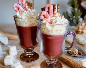 May include: Two clear glass mugs filled with a red beverage, topped with whipped cream, candy canes, and chocolate. The mugs sit on a wooden surface with marshmallows and cinnamon sticks.