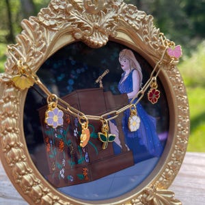 K&ouml;nnte beinhalten: Ein goldfarbener Bilderrahmen mit einer Zeichnung einer Frau in einem blauen Kleid an einem Rednerpult. Eine goldene Halskette mit Blumenanh&auml;ngern ist &uuml;ber dem Rahmen drapiert. Der Rahmen hat ein verziertes, dekoratives Design.