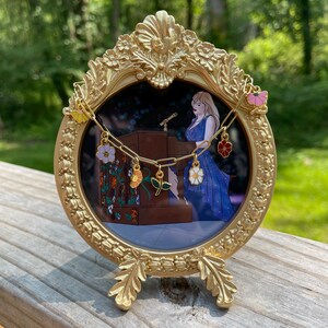 K&ouml;nnte beinhalten: Ein goldfarbener, verzierter Bilderrahmen mit runder &Ouml;ffnung, der eine Konzertszenen zeigt. Eine goldene Kette mit bunten Blumenanh&auml;ngern h&auml;ngt &uuml;ber dem Rahmen. Der Rahmen steht auf einer Holzoberfl&auml;che vor einem verschwommenen gr&uuml;nen Hintergrund.
