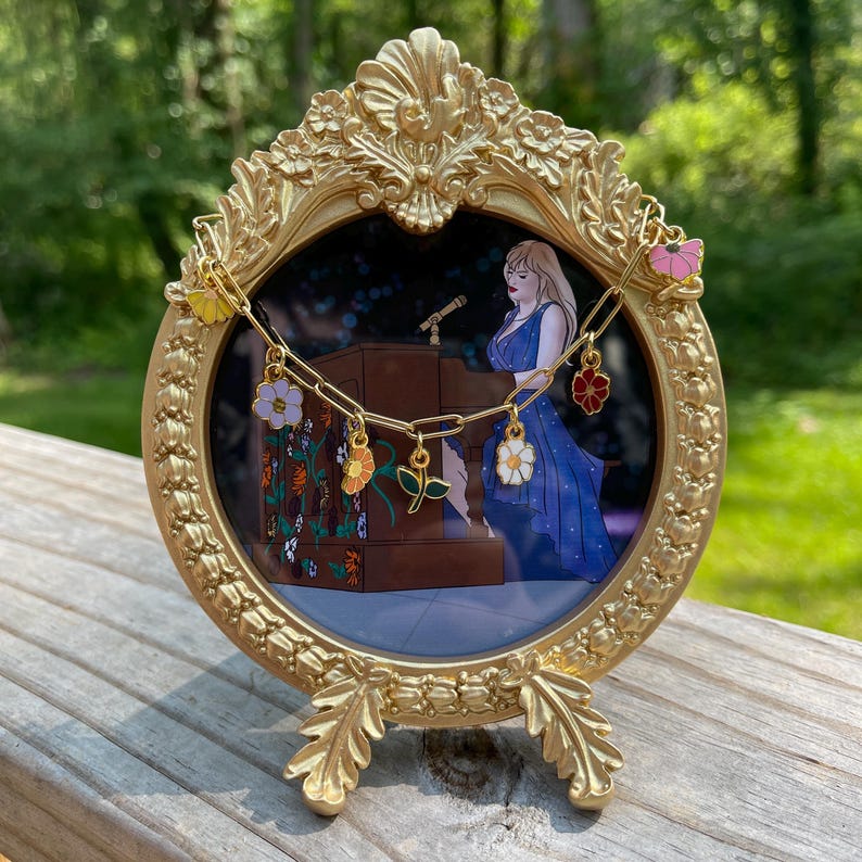K&ouml;nnte beinhalten: Ein goldfarbener, verzierter Bilderrahmen mit runder &Ouml;ffnung, der eine Konzertszenen zeigt. Eine goldene Kette mit bunten Blumenanh&auml;ngern h&auml;ngt &uuml;ber dem Rahmen. Der Rahmen steht auf einer Holzoberfl&auml;che vor einem verschwommenen gr&uuml;nen Hintergrund.