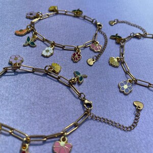 K&ouml;nnte beinhalten: Goldfarbene Charm-Armb&auml;nder mit bunten Blumen- und Blattanh&auml;ngern. Die Armb&auml;nder haben ein Kettendesign und werden auf einer lila Oberfl&auml;che pr&auml;sentiert. Die Charms umfassen Blumen in Rosa-, Gelb-, Wei&szlig;- und Lilat&ouml;nen.