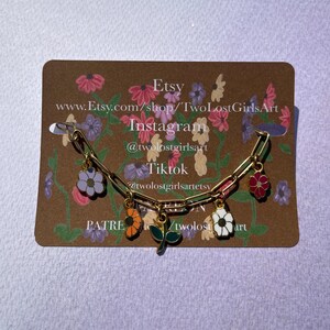 K&ouml;nnte beinhalten: Ein goldfarbenes Kettenarmband mit bunten Emaille-Blumenanh&auml;ngern. Die Anh&auml;nger umfassen eine lila, orange, wei&szlig;e und rote Blume sowie ein gr&uuml;nes Blatt. Das Armband wird auf einer floralen Karte mit Etsy-Shop-Informationen pr&auml;sentiert.