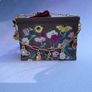 K&ouml;nnte beinhalten: Eine braune, rechteckige Handtasche mit einem Blumendesign mit Sonnenblumen, Rosen und anderen Blumen in Rosa, Gelb und Wei&szlig;. Die Tasche hat einen goldfarbenen Kettenriemen mit bunten Blumenanh&auml;ngern und einen bordeauxroten Samtgriff.