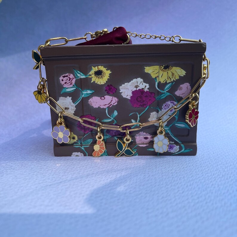 K&ouml;nnte beinhalten: Eine braune, rechteckige Handtasche mit einem Blumendesign mit Sonnenblumen, Rosen und anderen Blumen in Rosa, Gelb und Wei&szlig;. Die Tasche hat einen goldfarbenen Kettenriemen mit bunten Blumenanh&auml;ngern und einen bordeauxroten Samtgriff.