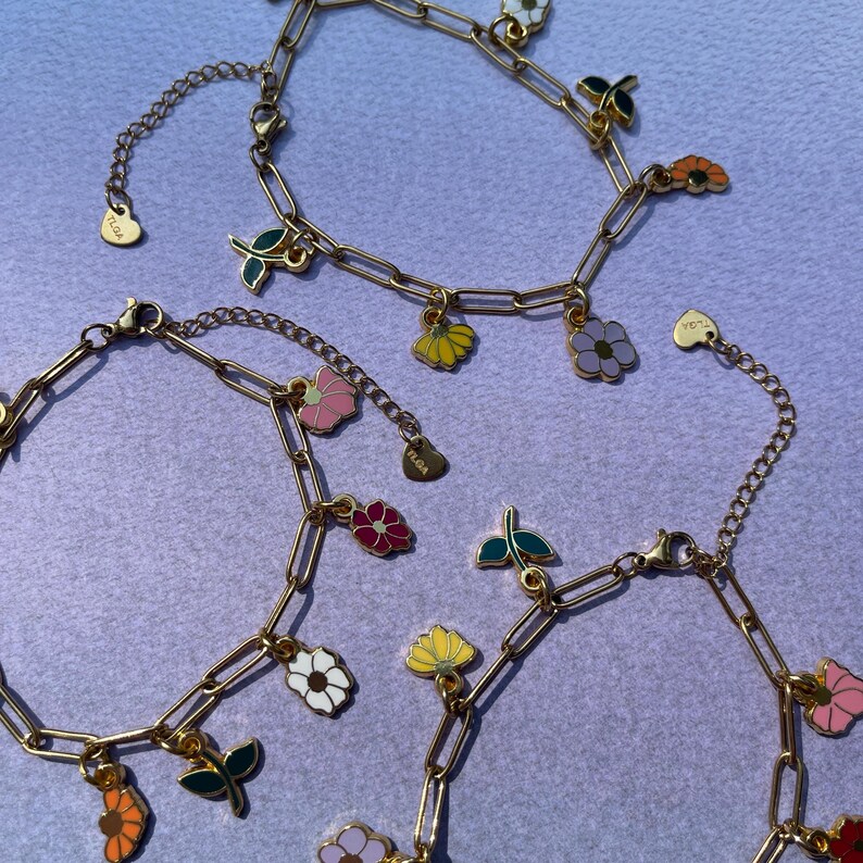 K&ouml;nnte beinhalten: Goldfarbene Kettenarmb&auml;nder mit bunten Emaille-Blumenanh&auml;ngern. Die Anh&auml;nger zeigen verschiedene Blumenmotive in Rosa, Gelb, Wei&szlig; und Lila sowie gr&uuml;ne Bl&auml;tter. Jedes Armband hat einen Karabinerverschluss.