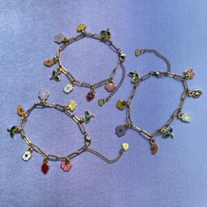 K&ouml;nnte beinhalten: Drei goldfarbene Kettenarmb&auml;nder mit bunten Blumenanh&auml;ngern. Die Anh&auml;nger umfassen verschiedene Blumendesigns in Rosa-, Gelb-, Orange-, Rot- und Wei&szlig;t&ouml;nen mit gr&uuml;nen Bl&auml;ttern. Die Armb&auml;nder werden auf einer hellvioletten Oberfl&auml;che pr&auml;sentiert.