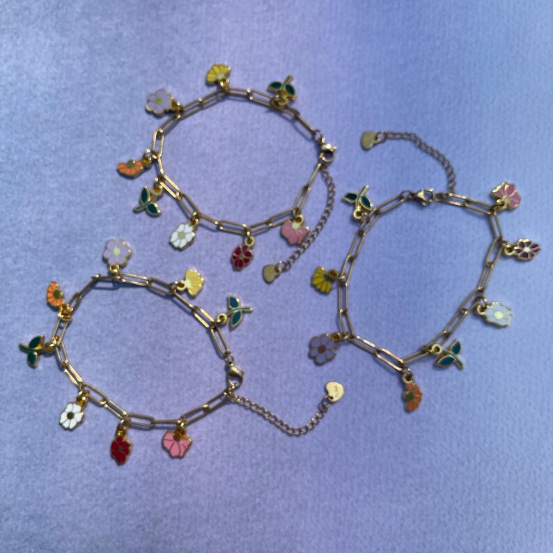 K&ouml;nnte beinhalten: Drei goldfarbene Kettenarmb&auml;nder mit bunten Blumenanh&auml;ngern. Die Anh&auml;nger umfassen verschiedene Blumendesigns in Rosa-, Gelb-, Orange-, Rot- und Wei&szlig;t&ouml;nen mit gr&uuml;nen Bl&auml;ttern. Die Armb&auml;nder werden auf einer hellvioletten Oberfl&auml;che pr&auml;sentiert.