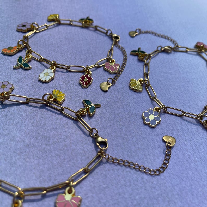 K&ouml;nnte beinhalten: Goldfarbene Kettenarmb&auml;nder mit bunten Emaille-Blumenanh&auml;ngern. Die Anh&auml;nger zeigen verschiedene Blumenmuster in Rosa, Gelb, Wei&szlig; und Lila. Die Armb&auml;nder werden auf einer hellvioletten Oberfl&auml;che pr&auml;sentiert.