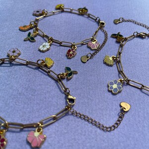 K&ouml;nnte beinhalten: Goldfarbene Kettenarmb&auml;nder mit bunten Emaille-Blumenanh&auml;ngern. Die Anh&auml;nger zeigen verschiedene Blumenmotive in Rosa, Gelb, Wei&szlig; und Lila, mit gr&uuml;nen Bl&auml;ttern. Die Armb&auml;nder sind auf einer lavendelfarbenen Oberfl&auml;che pr&auml;sentiert.