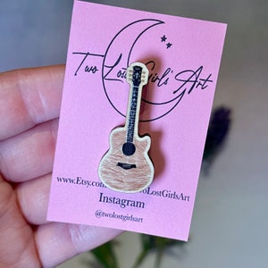 May include: A wooden guitar pin with a natural wood finish and black details. The pin is displayed on a pink card with the text "Two Lost Girls Art" and website information.
