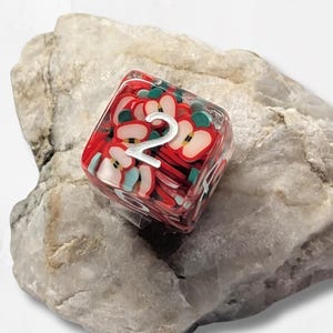 Könnte beinhalten: Ein transparenter, würfelförmiger Spielwürfel mit einem in das Harz eingebetteten Muster aus roten und weißen Äpfeln. Die Zahl Zwei ist in einer großen, silberfarbenen Schrift auf der Oberseite des Würfels zu sehen, der auf einem rauen Stein liegt.