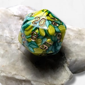 Puede incluir: Un dado de veinte caras con un diseño único. El dado presenta un patrón de formas amarillas y verdes en forma de maíz sobre un fondo verde azulado. Los números son de color dorado.