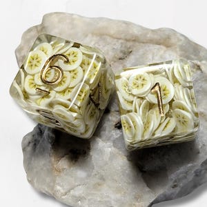 May include: Two dice with a clear resin exterior, filled with banana slice designs. The dice have gold-colored numbers, with one die showing the number 6 and the other showing the number 1. The dice are resting on a light gray stone.