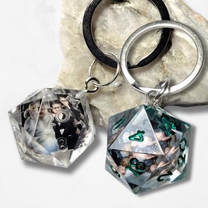 Op de afbeelding: Twee doorzichtige, veelzijdige dobbelsteen sleutelhangers. Eén dobbelsteen heeft een zwart-wit foto binnenin, de andere heeft groene accenten en cijfers. Beide zijn bevestigd aan metalen sleutelringen, één zwart en één zilver.