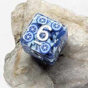 Könnte beinhalten: Ein sechsseitiger Würfel mit einem Körper aus durchscheinendem blauem Harz, in den kleine, kreisförmige Blumenmuster in Blau- und Weißtönen eingebettet sind. Die Zahl sechs ist auf der Oberseite in einer silberfarbenen Schrift.