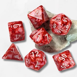 May include: A collection of seven red and white marbled polyhedral dice, ideal for role-playing games. The dice are displayed on a light grey rock. Each die shows a different number. The dice are made of a translucent material.