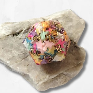 May include: A multi-colored, twenty-sided die filled with star-shaped confetti and gold-colored numbers. The die is resting on a light gray, textured stone. The numbers are in a gold color.