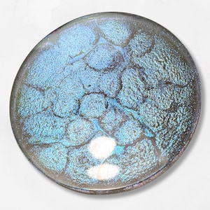 Könnte beinhalten: Ein rundes, irisierendes Glasobjekt mit einer strukturierten, blau-türkisfarbenen Oberfläche. Das Glas hat einen dichroitischen Effekt, der mehrere Farben reflektiert. Das Objekt ist vor einem weißen Hintergrund platziert, der seine einzigartige Farbe und sein Design hervorhebt.