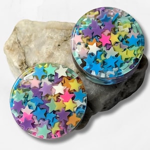 May include: Two clear, round plugs filled with colorful star-shaped confetti. The plugs are set on a light gray rock against a white background. The stars are various colors, including pink, yellow, blue, and purple.