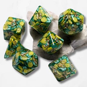 Puede incluir: Juego de siete dados poliédricos con un patrón amarillo y verde similar al maíz. Los dados tienen números dorados y están dispuestos sobre una superficie blanca. Los dados son para juegos de mesa.