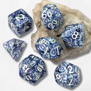 Könnte beinhalten: Ein Set aus sieben polyedrischen Würfeln mit einem blau-weißen, blumenartigen Millefiori-Muster und weißen Zahlen. Das Set enthält einen W4, W6, W8, W10, W12, W20 und einen Prozentwürfel, die auf einem strukturierten, hellen Stein liegen.