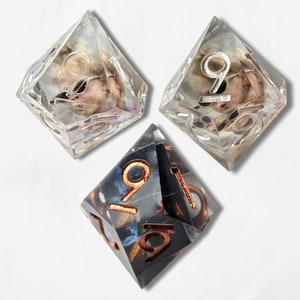 May include: Three eight-sided dice arranged in a triangular formation. Two dice are clear with internal beige and brown inclusions and silver-coloured numbers. The third die is dark grey with internal brown swirls and copper-coloured numbers.