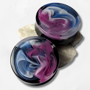 May include: Two round ear plugs with a swirling design in shades of blue, white, and magenta. The plugs are set against a neutral stone background, showcasing the colorful, marbled effect of the resin.