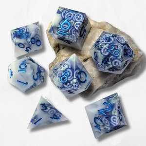 Könnte beinhalten: Satz von sechs weißen Würfeln mit blauen und silbernen Akzenten. Die Würfel haben ein wirbelndes Muster und sind von 1 bis 6 nummeriert. Die Würfel befinden sich auf einer weißen Oberfläche.