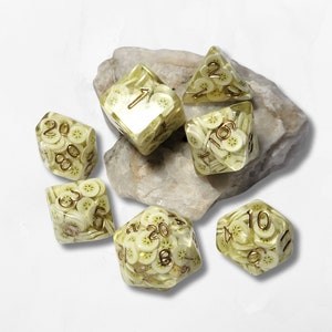 May include: Set of six dice with a banana slice design. The dice are clear with a yellow banana slice pattern and gold numbers. The dice are on a rock.