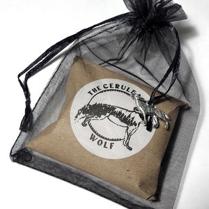 May include: A silver wolf pendant necklace in a black mesh bag with a brown paper insert. The insert has a white circle with the text "The Cerule Wolf" and a stylized wolf illustration.