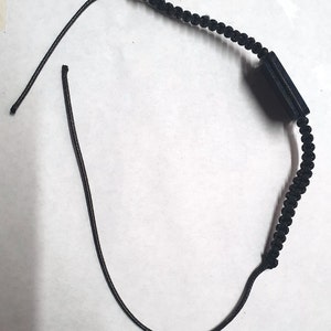 May include: A black cord bracelet with a black rectangular charm. The cord is braided and the charm is smooth.