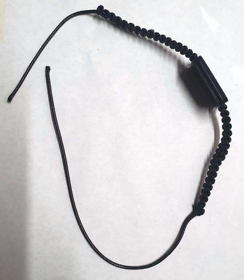 May include: A black cord bracelet with a black rectangular charm. The cord is braided and the charm is smooth.