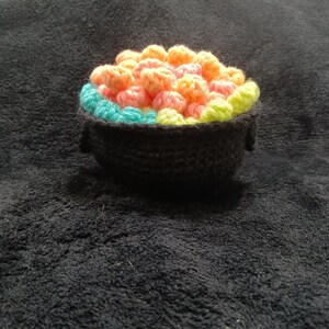 May include: A crocheted pot filled with colorful balls, resembling a pot of gold. The pot is black with a green and blue rim.